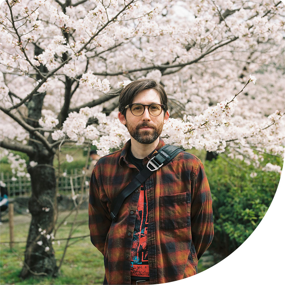 Michael Harper, a white person with brown hair, stands in front of a cherry blossom tree wearing a brown plaid flannel.