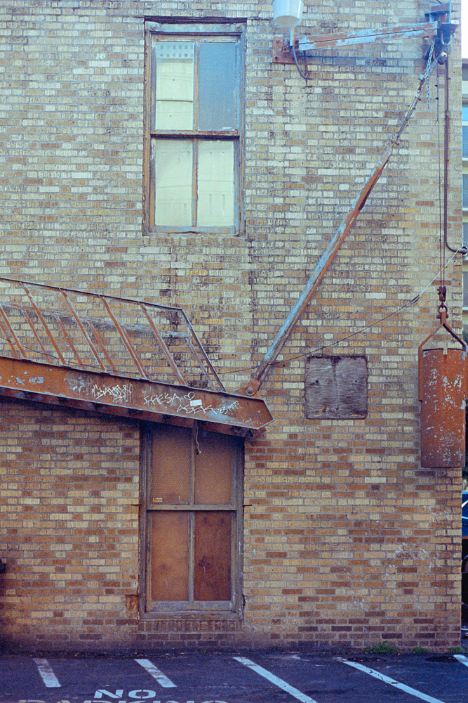 A dirty, red brick wall in a parking lot. A rusted fire escape hangs on the side.